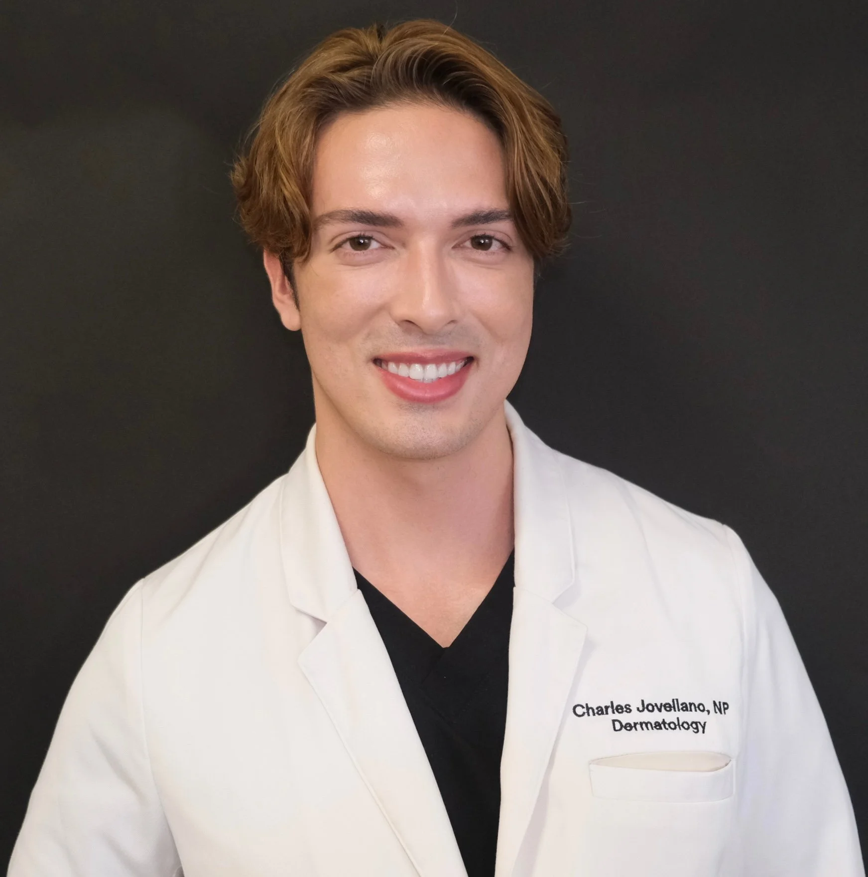 Charles Jovellano, NP in Dermatology, wearing white lab coat over black shirt, smiling at camera